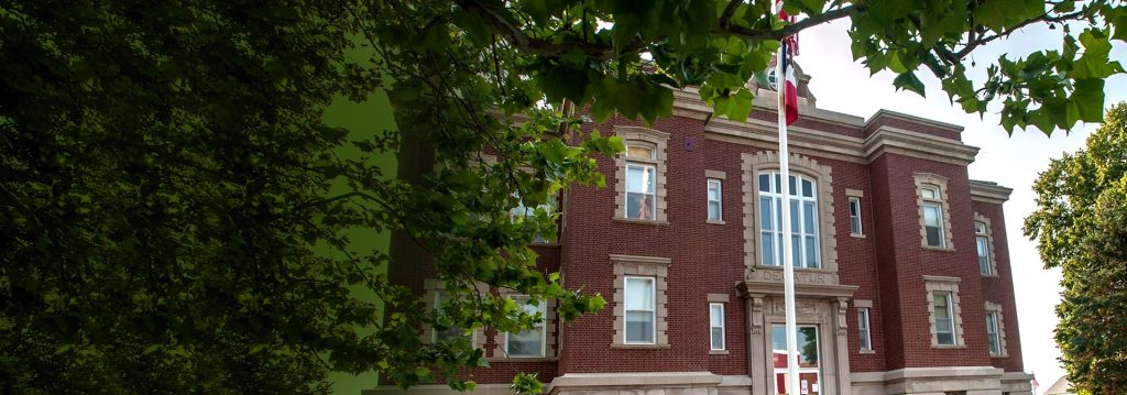 A large, historic red brick building with white trim and tall windows—home to the Leon Chamber of Commerce—is partially framed by leafy green trees, with a flagpole in front and a bright blue sky in the background.