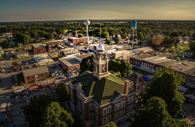 leon sky view Aerial view of a small town with a historic courthouse in the foreground, surrounded by trees, and a downtown area with buildings, water towers, and people gathering on the streets. The landscape is green and stretches into the distance.